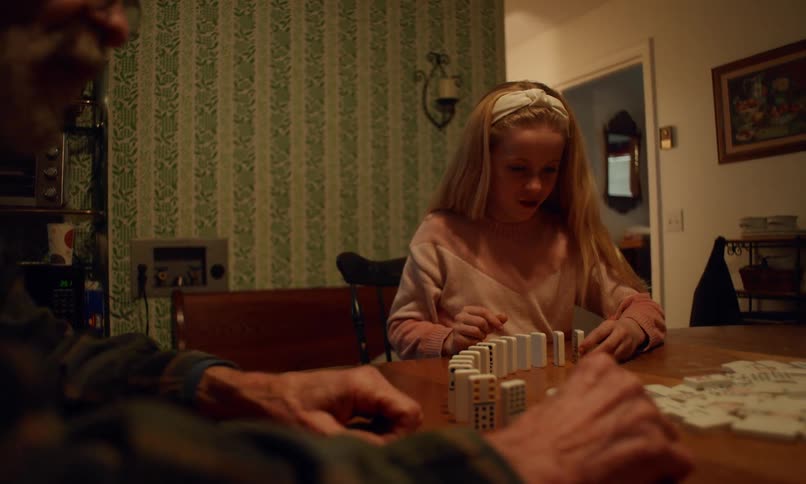 A young girl in a pink sweater is carefully setting up a line of dominoes on a wooden table while focusing on her game