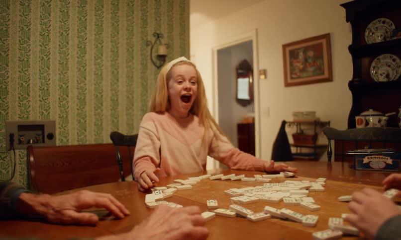 An elderly man wearing a beanie and glasses is sitting at a wooden table watching as a young girl in a pink sweater reacts in surprise while the dominoes she set up start to fall