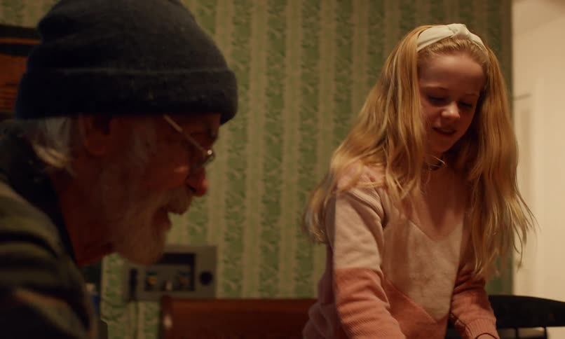 An elderly man with a gray beard wearing a beanie and glasses sits at a wooden table smiling warmly while a young boy in the foreground laughs and a blonde girl in the background watches with excitement in a cozy kitchen
