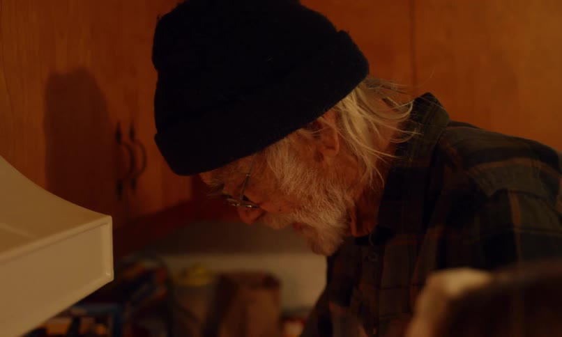 Elderly man in cap and glasses looks down in a wooden kitchen lit by warm light