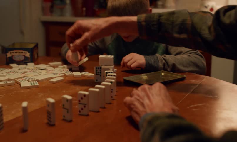 An intergenerational group plays dominoes around a wooden table in a casual kitchen setting