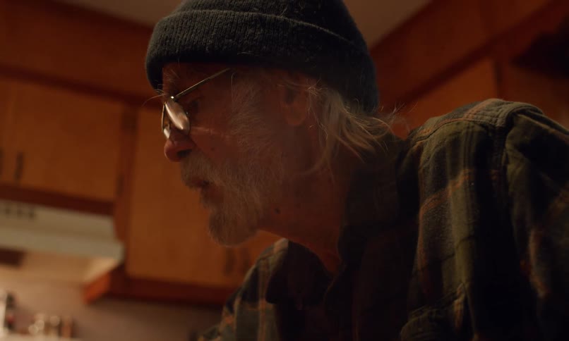 Elderly man in a cap and glasses looks intently at something out of frame in a kitchen