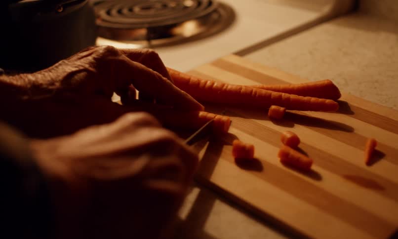 A pair of aged hands carefully chops fresh carrots on a wooden cutting board under the warm glow of the kitchen light