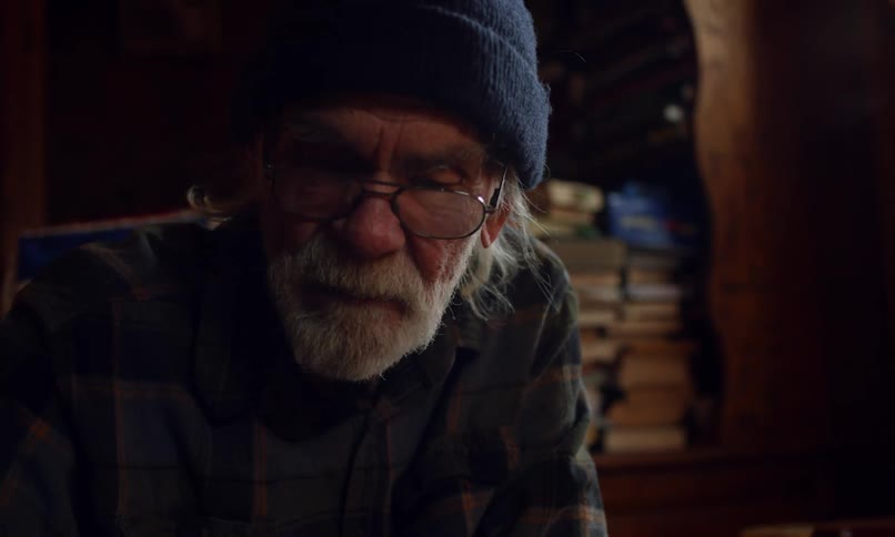 An elderly man with a white beard wearing a navy blue beanie and glasses engaged in a board game in a dimly lit cozy room filled with bookshelves stacked with books creating an intellectual and nostalgic atmosphere