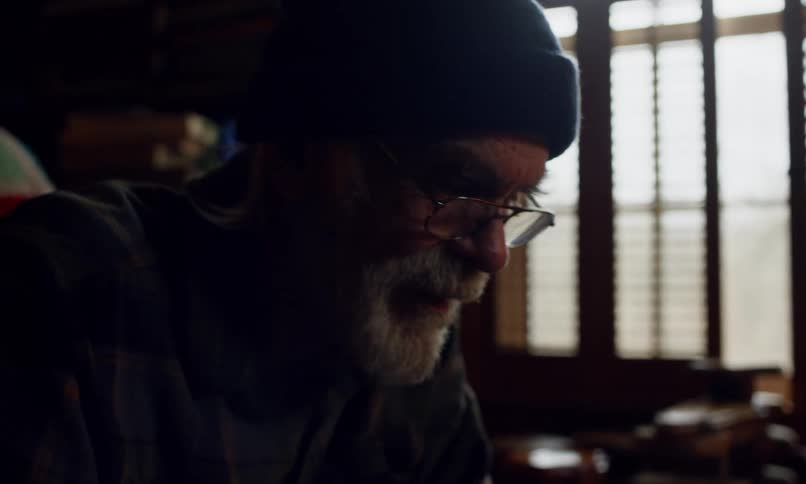 Elderly man with a gray beard and glasses wearing a blue beanie stands in a lowlit room
