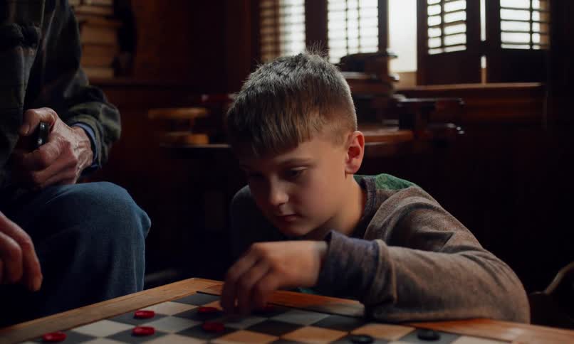 The face of a little boy who is playing checkers in a partially dark room with a window in the background
