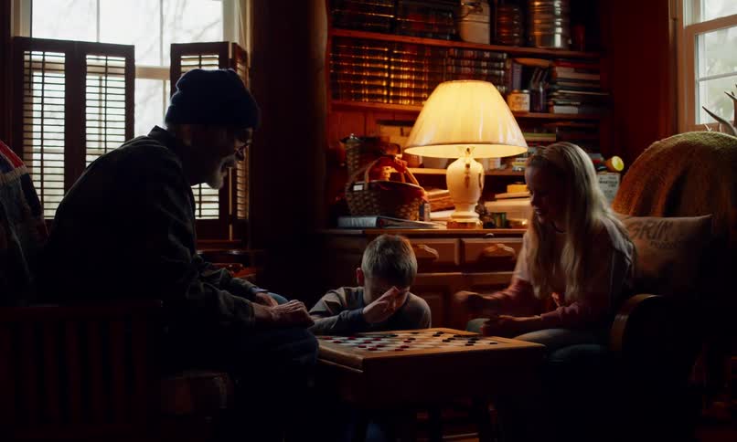 An elderly man a young boy and a young girl are silhouetted as they play checkers in a dimly lit wood paneled room