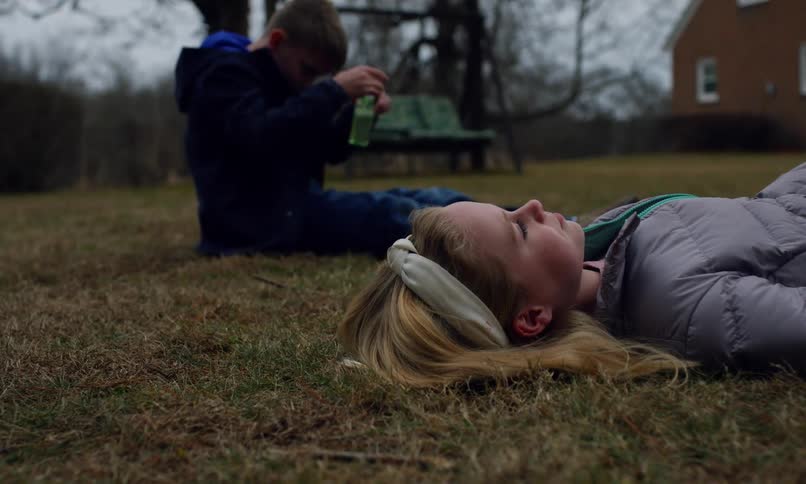 A girl lays on the grass looking at the sky while a boy is playing with a bottle of bubbles