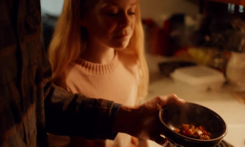 An adult is serving a bowl of stew to a blonde girl in a cozy kitchen at home