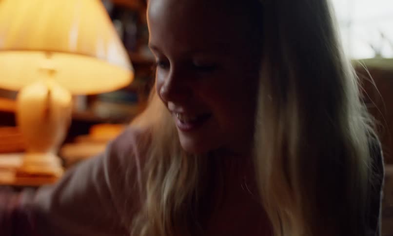 A young girl with a bright smile claps her hands joyfully sitting in a cozy room illuminated by the warm glow of a table lamp
