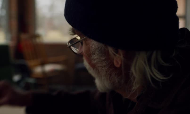 An elderly person with wrinkled hands is drawing on a large sheet of brown paper using crayons surrounded by scattered colorful crayons in a dimly lit cozy setting