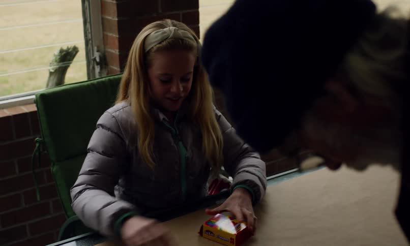 A young girl in a jacket happily opens a box of crayons while sitting at a table with two others