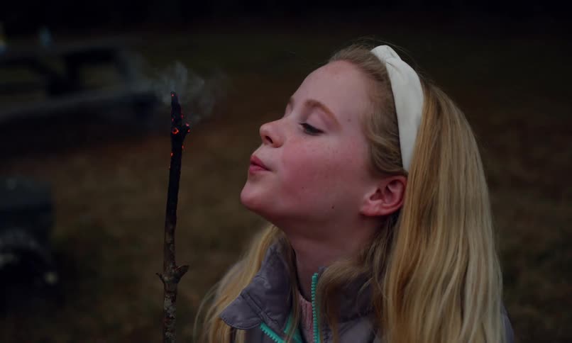 A blonde girl with a white headband is blowing on a burning stick trying to put the flame out