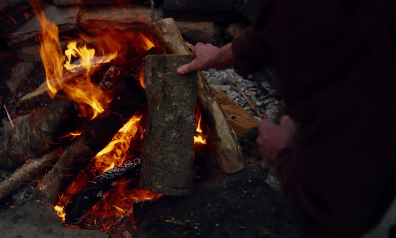 An elderly man places a log on a campfire in the countryside