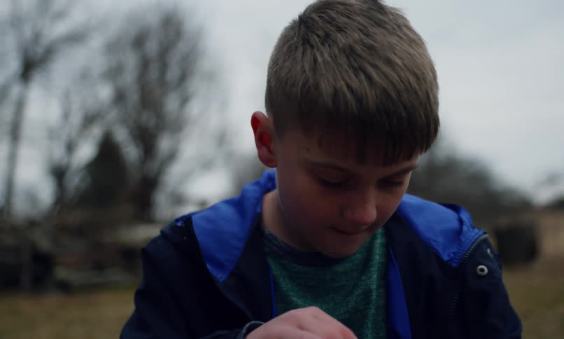 A young boy in a blue jacket blows air through a bubble wand his face full of concentration against a blurred outdoor background