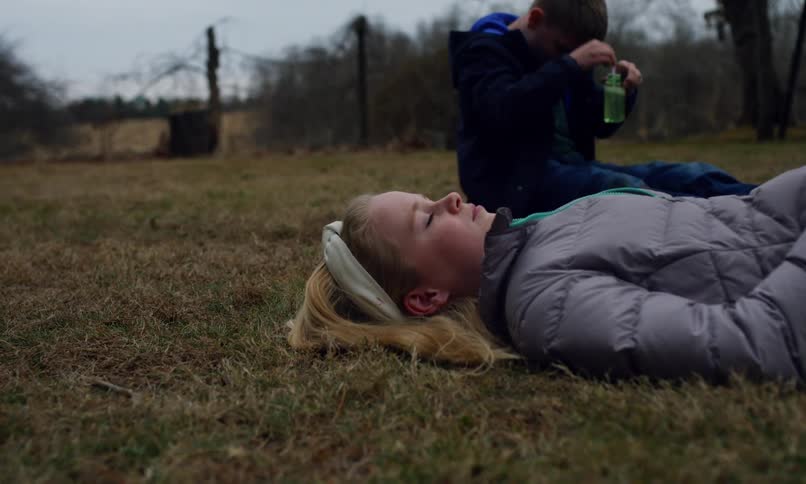 A girl in a gray jacket lies on the grass while a boy sits nearby holding a bubble wand and a bottle of bubble solution