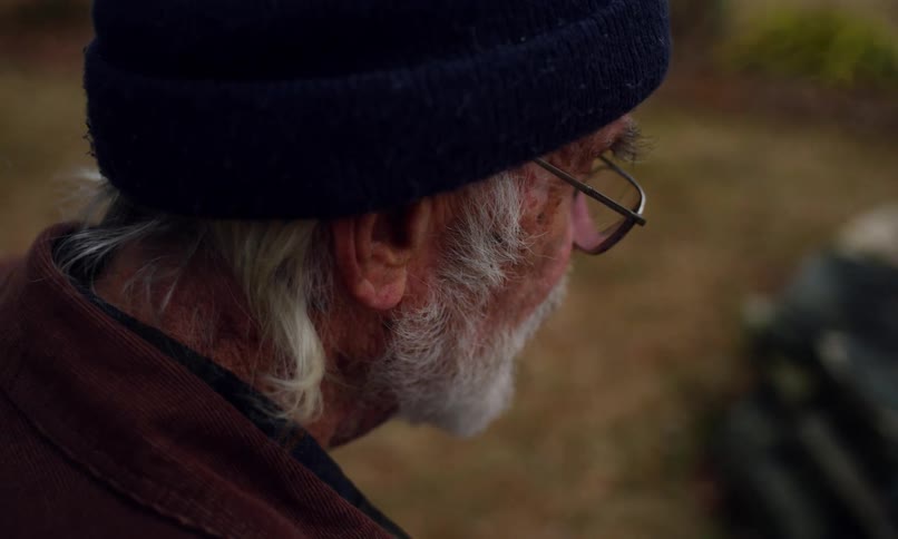An elderly man with white beard and hair sits outdoors listening to a girl