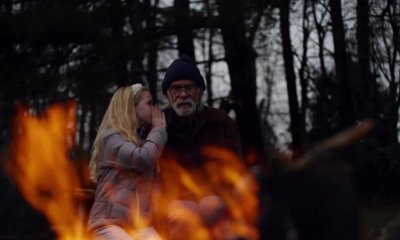 Little blonde girl sitting next to her grandfather at a campfire whispering in his ear