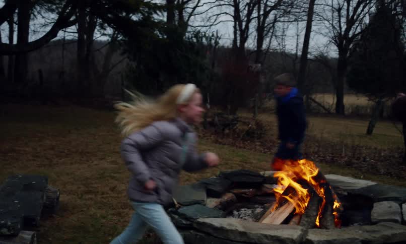 Amidst an autumnal campsite a grandfather and his two grandchildren a lively girl and boy are joyfully engaged in a playful game of chase as they excitedly run around a crackling bonfire in a field with stark bare trees capturing a heartwarming moment