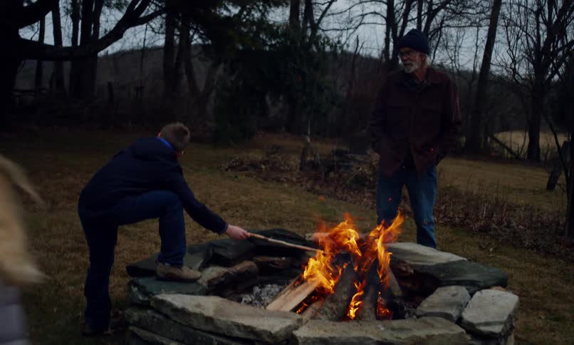 A young boy is tending to a campfire while an older man watches and a blonde girl is running around in the background