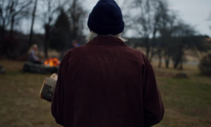 An elderly person carries logs to a stone fire pit with a burning fire in a grassy area while children play nearby