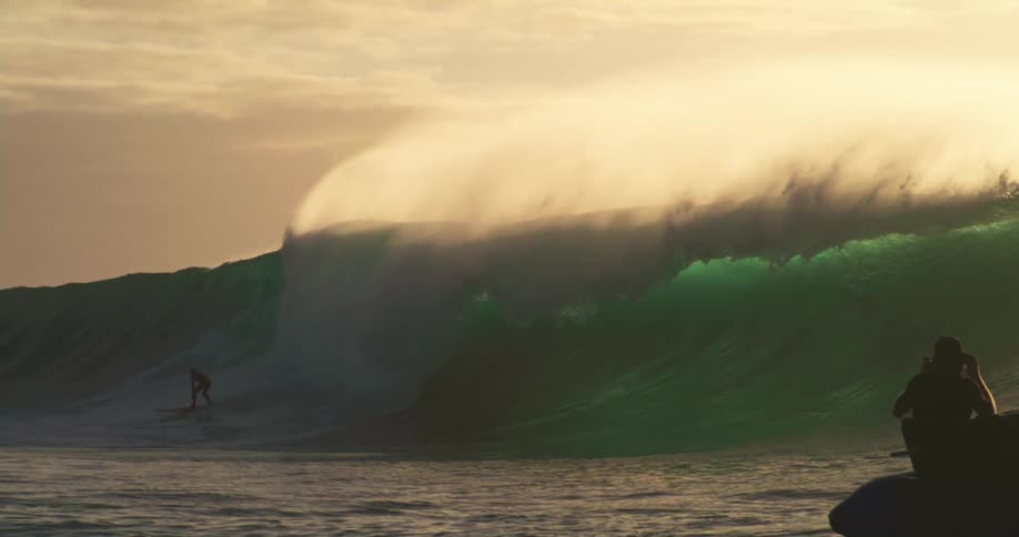Photographers on a boat capture a surfer riding a giant wave at sunset