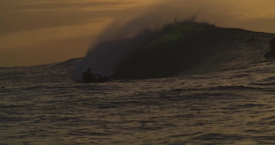 A sea wave breaking near some people who are on the jet ski on a morning with orange sky