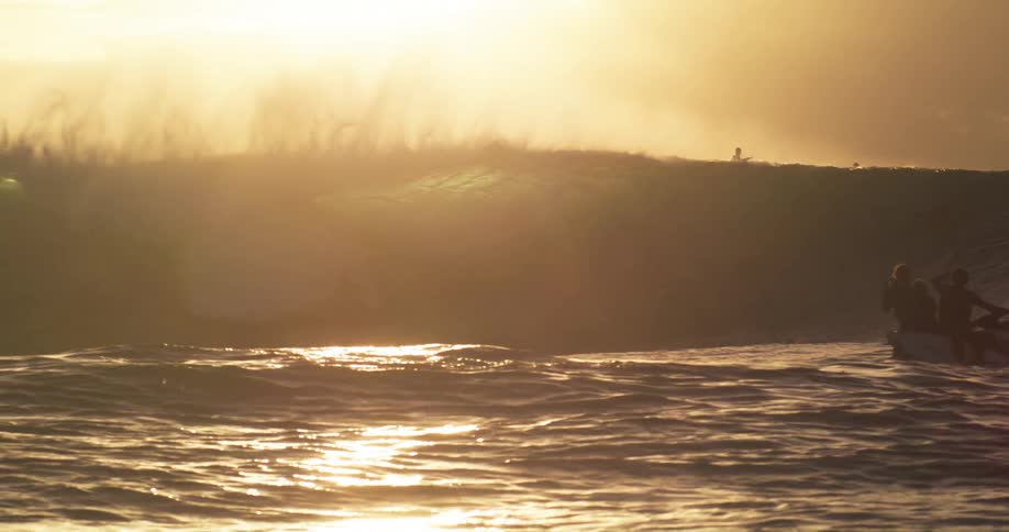 People ride a jet ski near the foot of a large wave while surfers sit on their boards near its top under a red sky