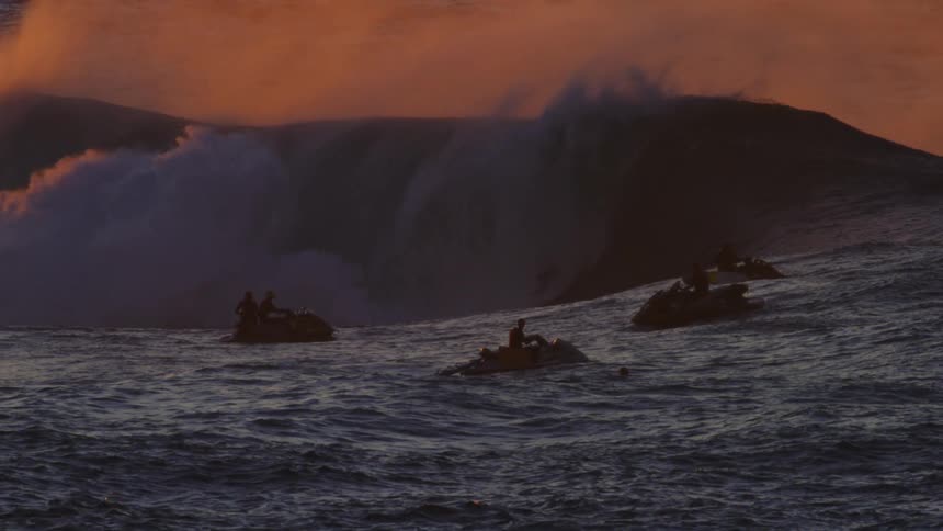 Silhouettes of people on jet skis following a surfer on a wave