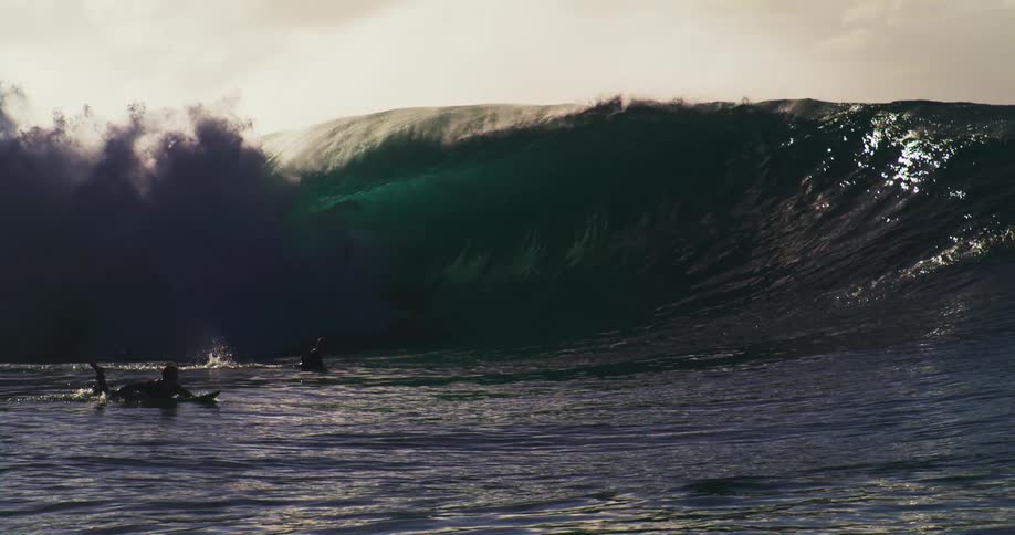 A huge wave breaking near some surfers who are paddling forward