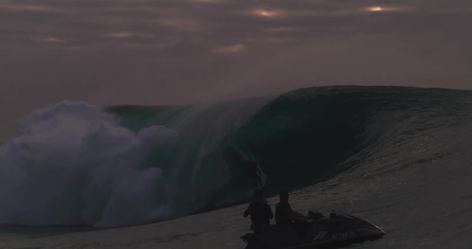 A surfer rides a large wave as two silhouetted figures on a jet ski watch from the foreground beneath a dusky sky