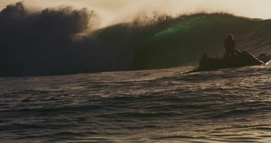 People on jet skis float in choppy ocean waters with a large wave cresting in the background and several surfers visible on the wave