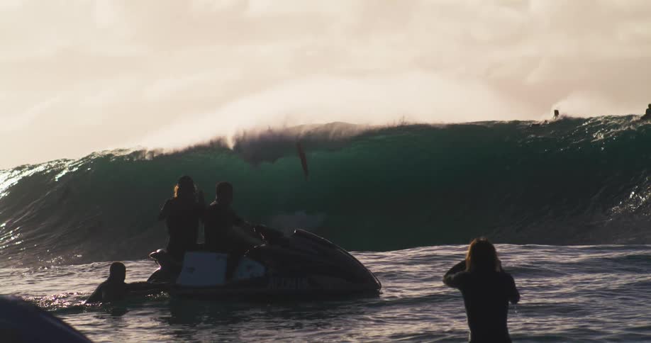 Some surfers are waiting for a big wave that is breaking to pass over them and in front there are some people watching and using jet ski