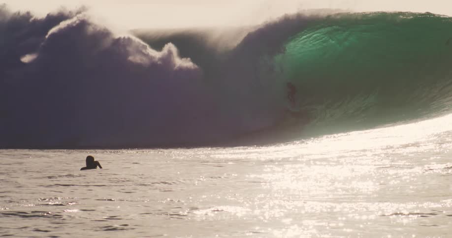 A big ocean wave is breaking next to some surfers and one of them is standing surfing on this wave