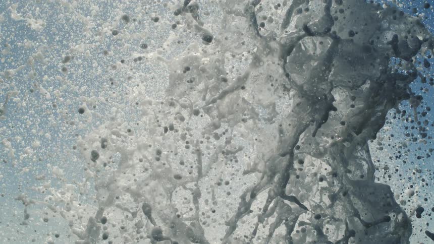 A closeup captures a burst of white foamy water splashing upwards against a light blue sky