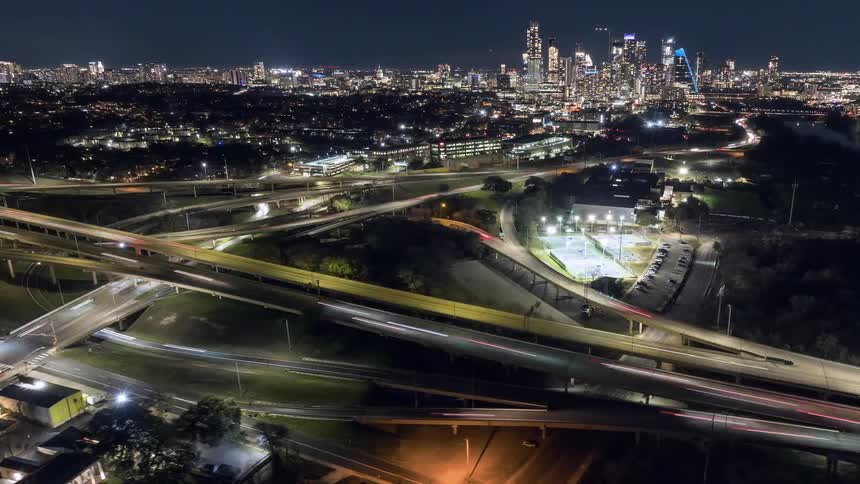 An elevated nighttime view shows a sprawling cityscape with illuminated skyscrapers in the distance segmented by a complex network of highways and roads with visible traffic
