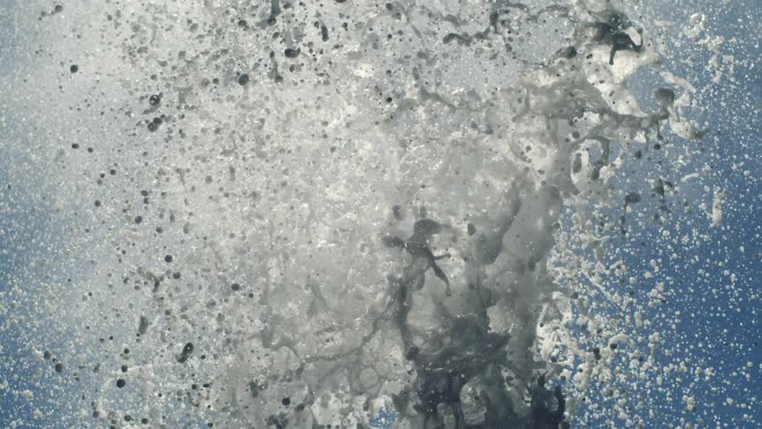 A closeup captures a burst of white water splashing upwards against a light blue sky