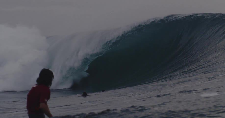 A huge wave is breaking in a tube shape next to some surfers who are watching