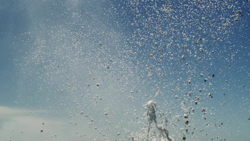 A closeup captures a burst of white water splashing upwards against a light blue sky