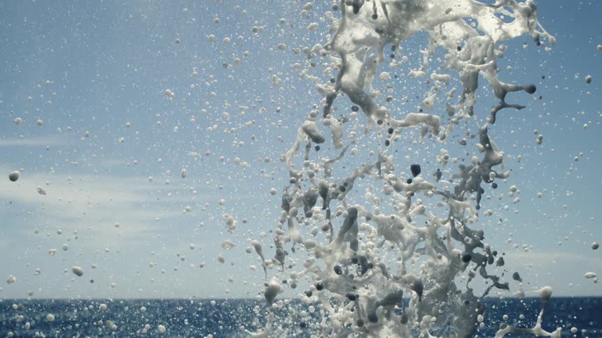 A closeup captures a burst of white water splashing upwards against a light blue sky and a dark blue horizon