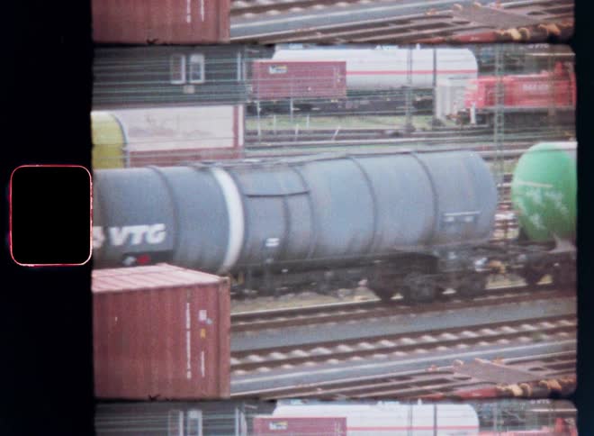 Evoking an 80s aesthetic with grainy filmlike skip effects an analog video captures a view from a passing train traversing a vast train yard filled with numerous tanker cars and containers on a complex network of tracks