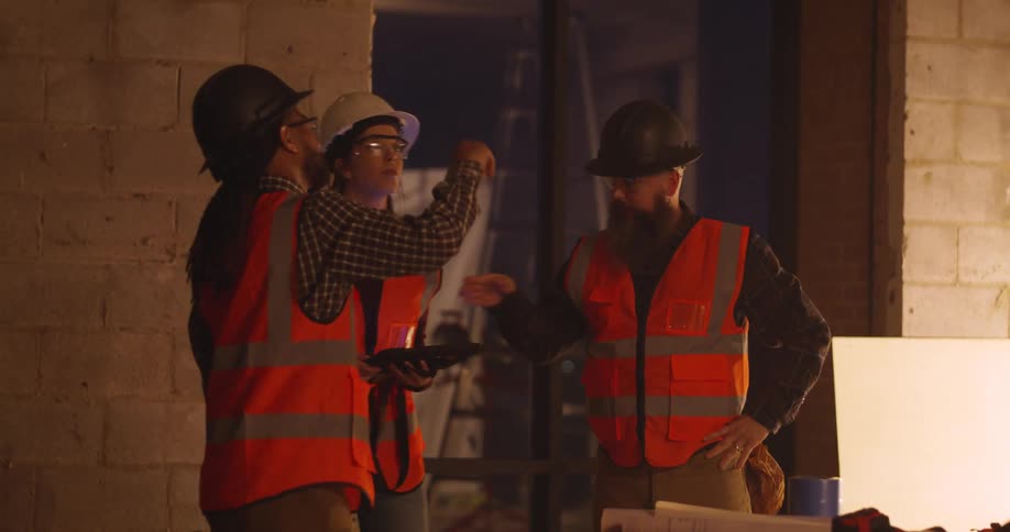 Three workers wearing hard hats, orange safety vests, and eye protection are standing at a job site while consulting a tablet and gesturing and pointing to spots in the building.