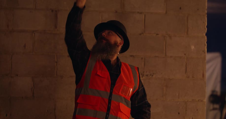 A bearded construction worker pointing something out while working at a construction site at night