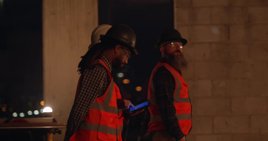 Construction workers wearing reflective vests and hard hats walk into a building site in the city at night carrying a tablet and stop to look around and discuss the project.