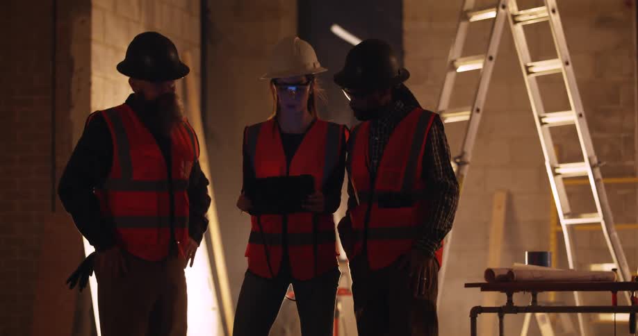 Three construction workers, a woman and two men, talk over what they're seeing on a tablet as they stand in front of a floodlight in a building under construction at night.
