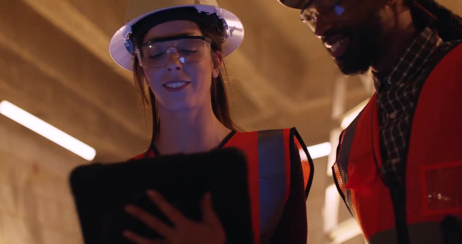 A man and a woman in an indoor construction site wearing hard hats and reflective vests consult a tablet and nod, then the woman leans to talk with another person to their right.