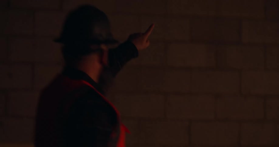 From a dark outdoor view of city lights, a bearded construction worker enters a brightly lit job site and uses clear hand signals to direct the flow of work and movement of other personnel within the busy workspace.