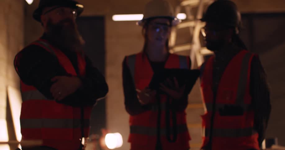 View panning from behind tools of three construction workers wearing hard hats and reflective vests who are reviewing information on a tablet at a construction site at night