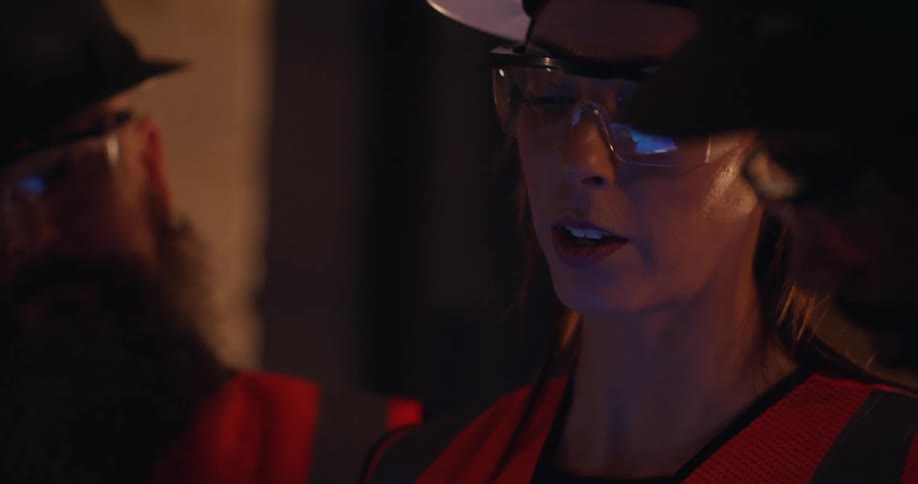 Close up of three engineers wearing protective gear and hard hats who are inspecting and discussing a building site over a tablet that's reflected in their safety glasses.