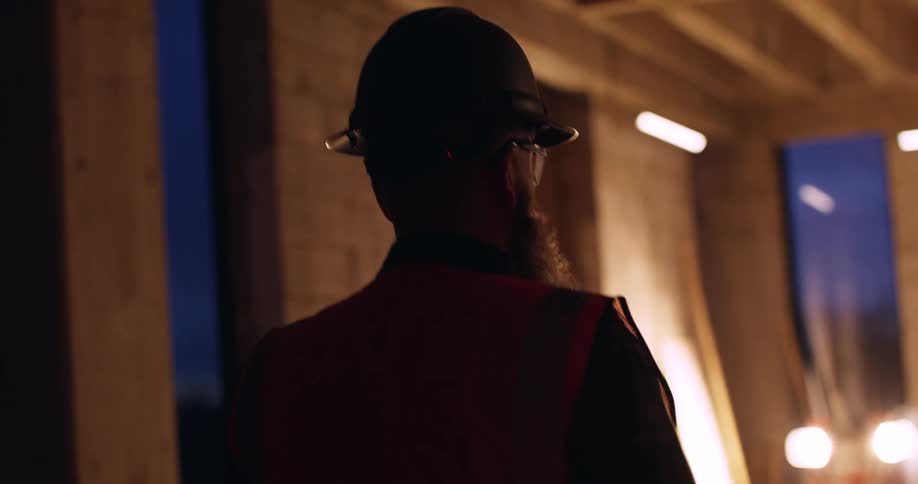 View from behind of a bearded construction worker wearing a hard hat and safety gear who is walking through an indoor building site at night and gesturing to different areas as he interacts with someone off camera.